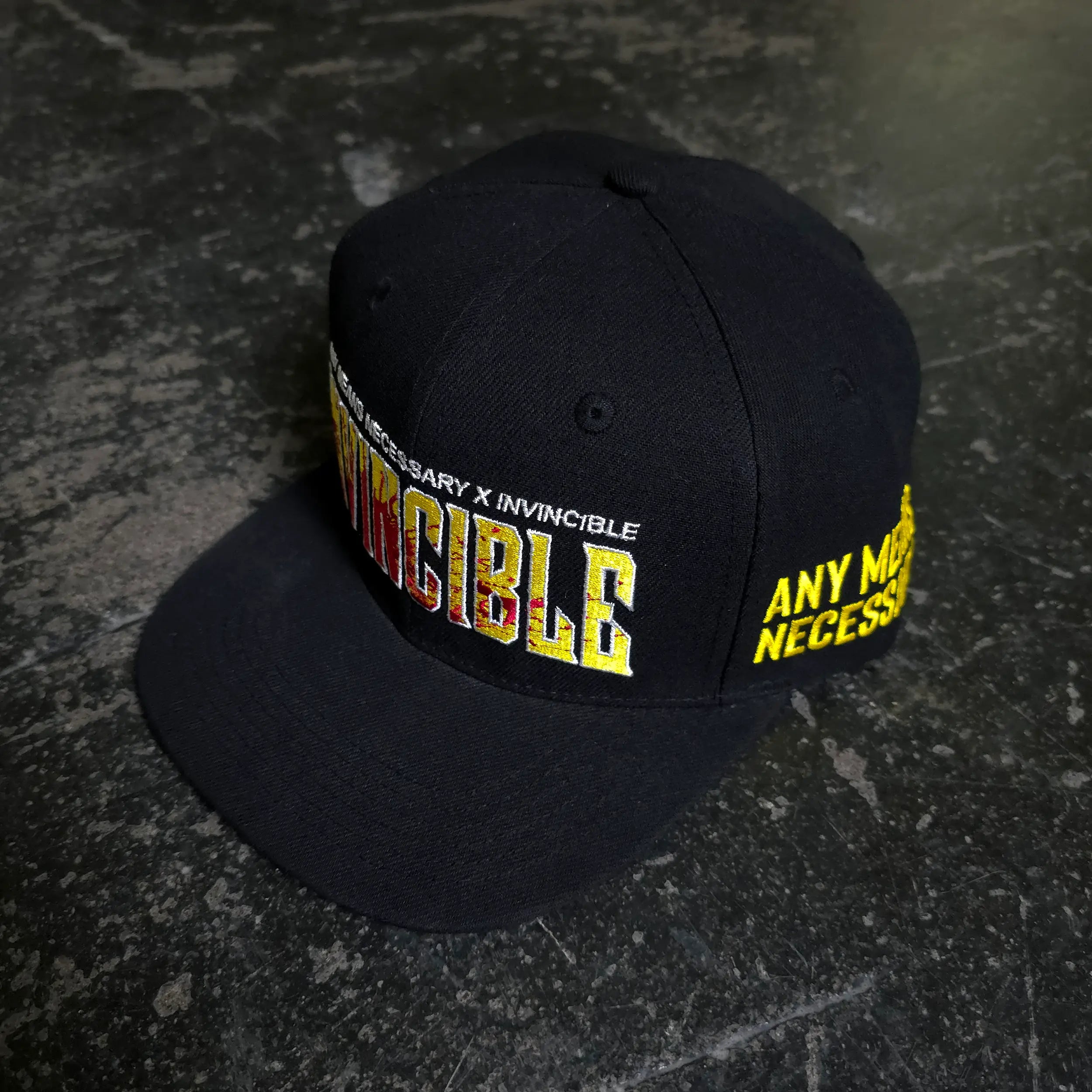 Black Invincible Splatter Hat by Any Means Necessary Clothing featuring bold paint splatter design and adjustable strap.
